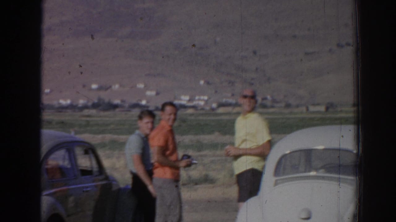 Three adults stand between two rounded cars in a rural roadside landscape.