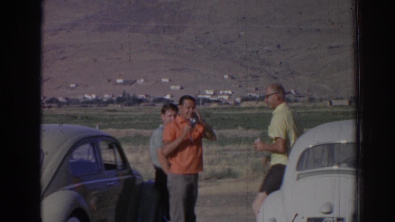Adults stand beside parked rounded cars while one person drinks from a bottle.