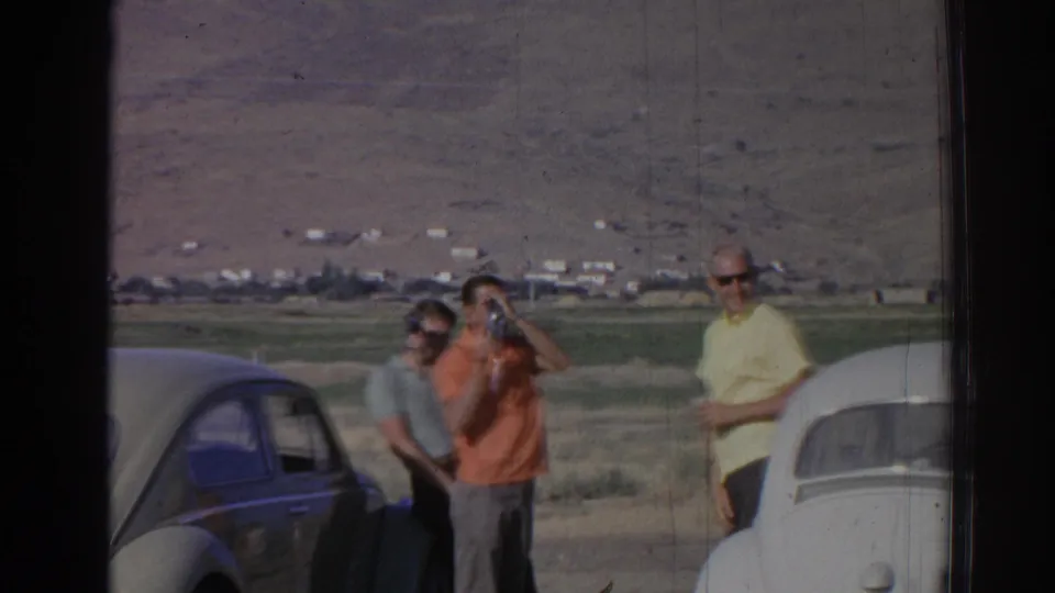 The 1962 Izmir home movie shows adults by two parked cars, including one person drinking while others stand in the rural landscape.