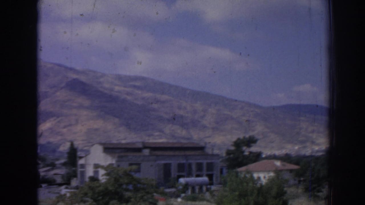 Window-framed view of a residence below rugged mountains under a partly cloudy sky.