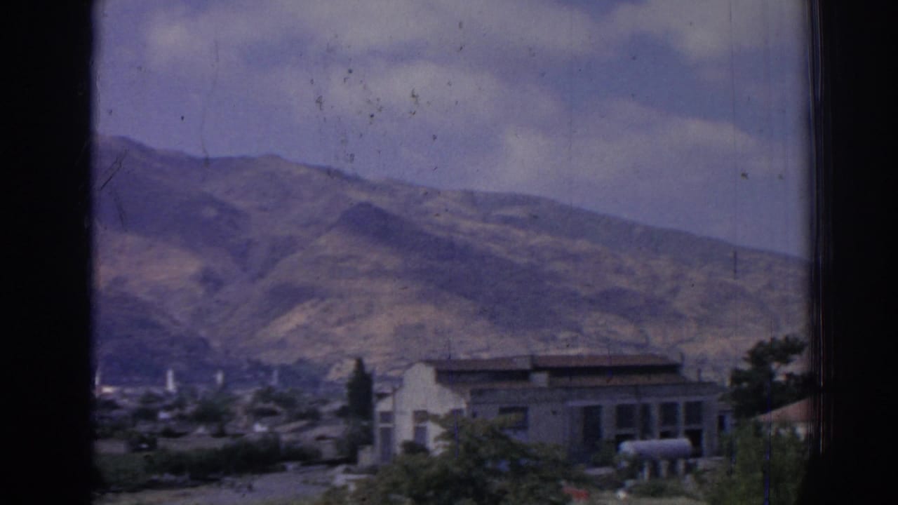Panned view through a window with mountains, a pale building, shrubs, and town structures.