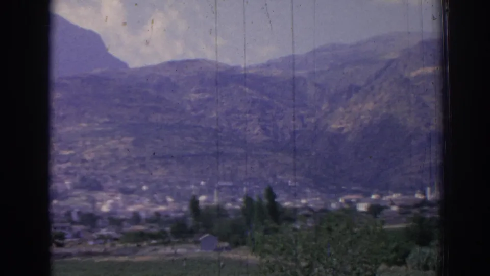 Vintage 8mm Izmir landscape with a distant town at the base of mountains, green fields, trees, and heavy dark edge framing.
