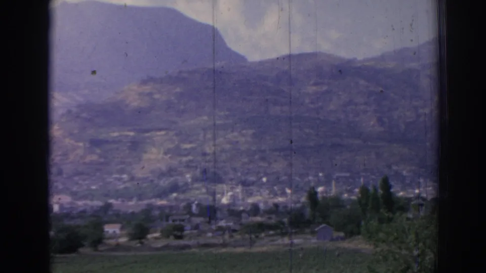 1962 Izmir view with low town buildings, slender minarets, trees, and purple-toned mountains under a pale sky.