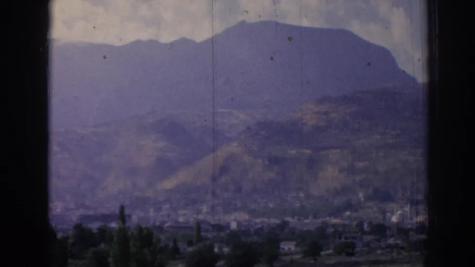 1962 8mm frame of Izmir from above, with a hazy mountain slope, valley buildings, foreground trees, and aged film texture.