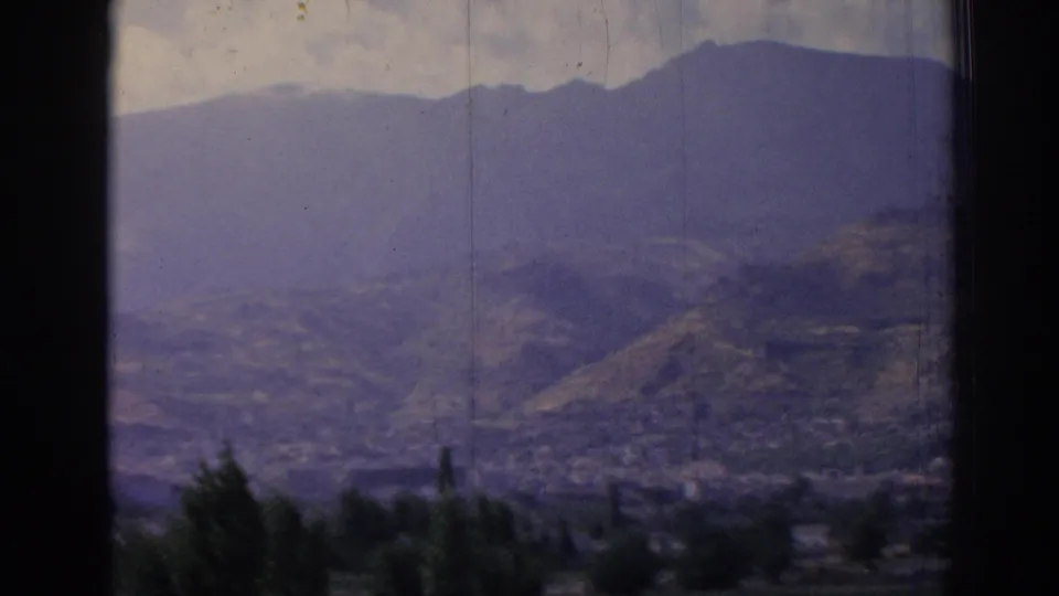 Late clip frame from the 1962 Izmir panorama, showing mountain ridges, a valley city, foreground foliage, and faded 8mm color.