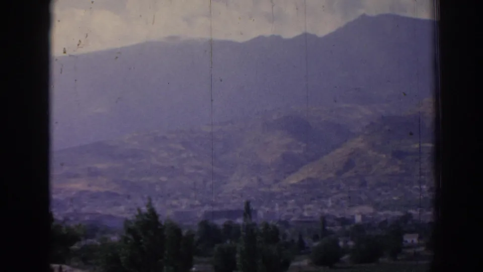 1962 8mm view from a window toward a hazy mountain valley in Izmir, Turkey, with foreground trees and distant settlement.