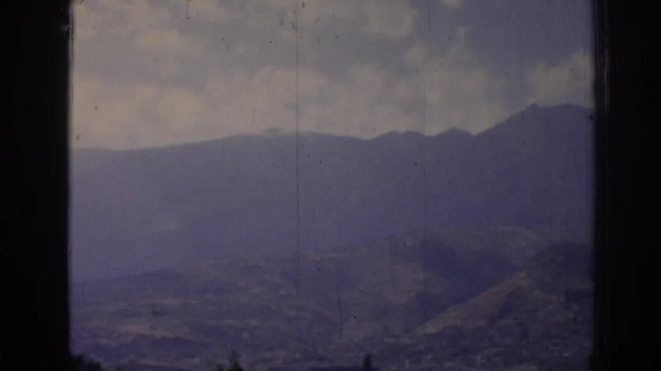 1962 8mm view near Izmir, Turkey, showing a distant mountain ridge, hazy hills, and clouds framed by a dark window edge.