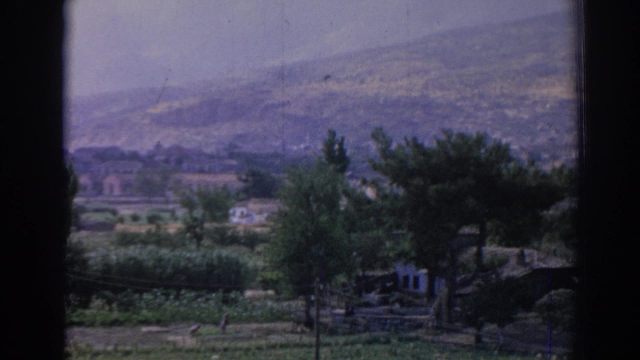 Window-framed rural view with trees, low buildings, open ground, and hazy hills.