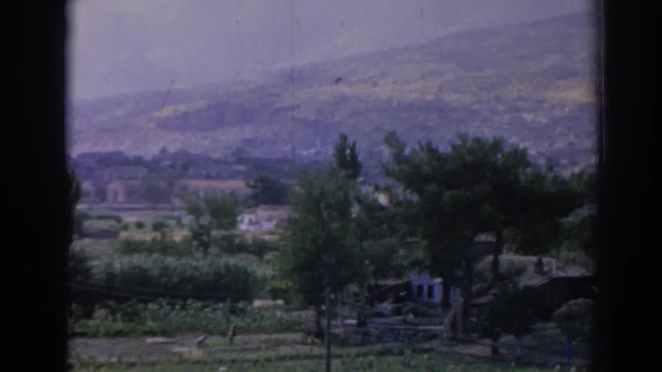 1962 8mm view in Izmir, Turkey, looking past dark window edges toward trees, low buildings, open ground, and distant hazy hills.