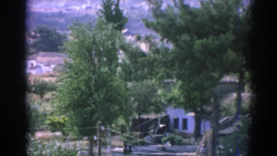 A 1962 Izmir home-movie frame with green trees, a small white building, utility lines, and dark vertical window edges.