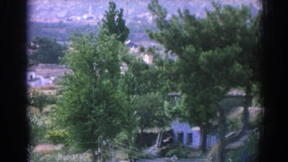 1962 Izmir-area home-movie still showing tall trees in front of a small white house, with low buildings and hazy hills beyond.