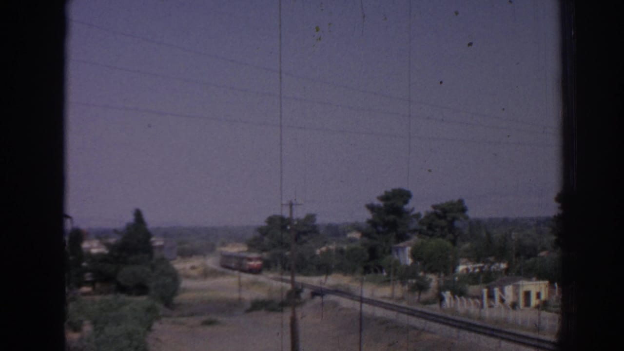 1962 Izmir home-movie frame showing railway tracks, distant train cars, trees, utility poles, and hazy landscape viewed through a dark window frame.