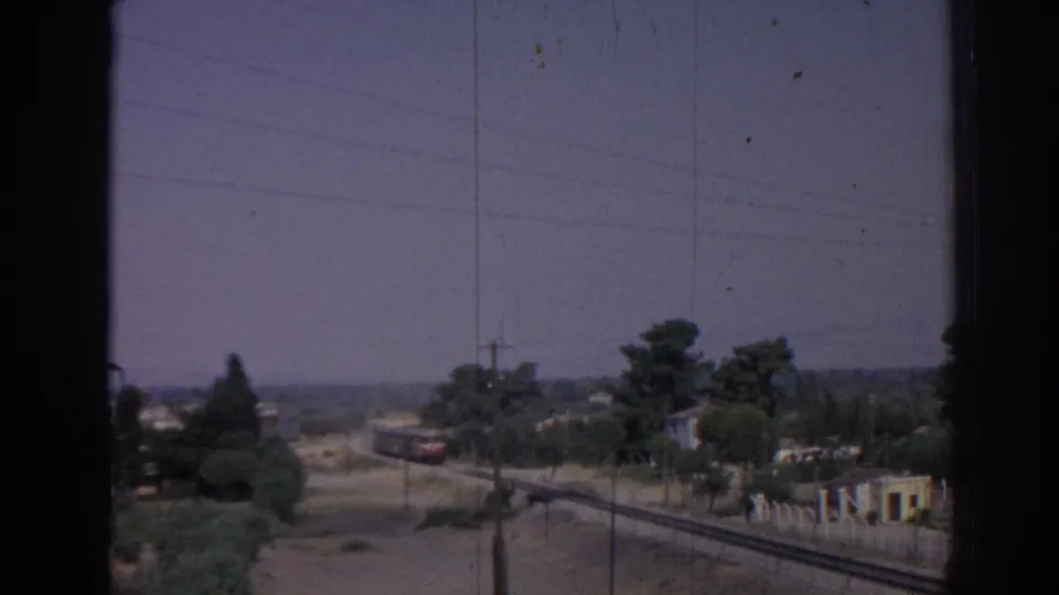 1962 Izmir home-movie frame showing railway tracks, distant train cars, trees, utility poles, and hazy landscape viewed through a dark window frame.