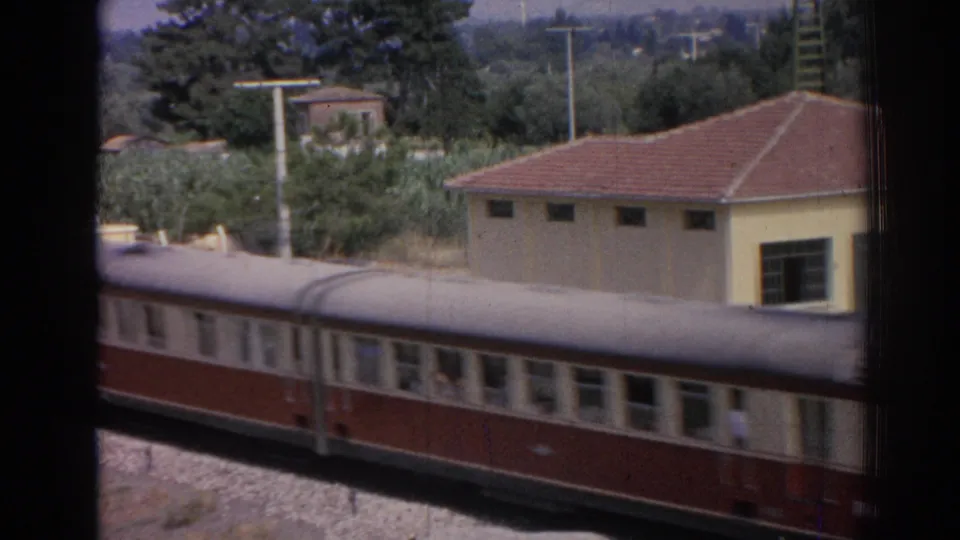A red-and-cream passenger train rolls past in the foreground, with a yellow station-side building, red roof tiles, trees, and utility poles behind it in Izmir.