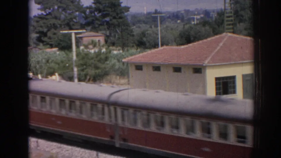 Later frame of the passing red-and-cream train, with passenger windows, a yellow building, red roof, trees, and hazy background visible from the elevated window view.