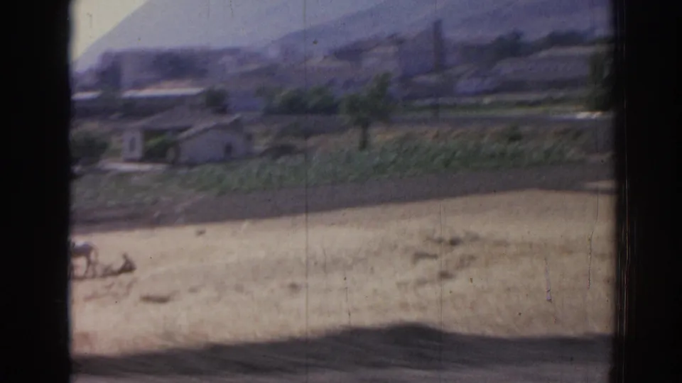 1962 Izmir-area countryside frame with a farmer and two horses working across a dry field, seen from a moving viewpoint.