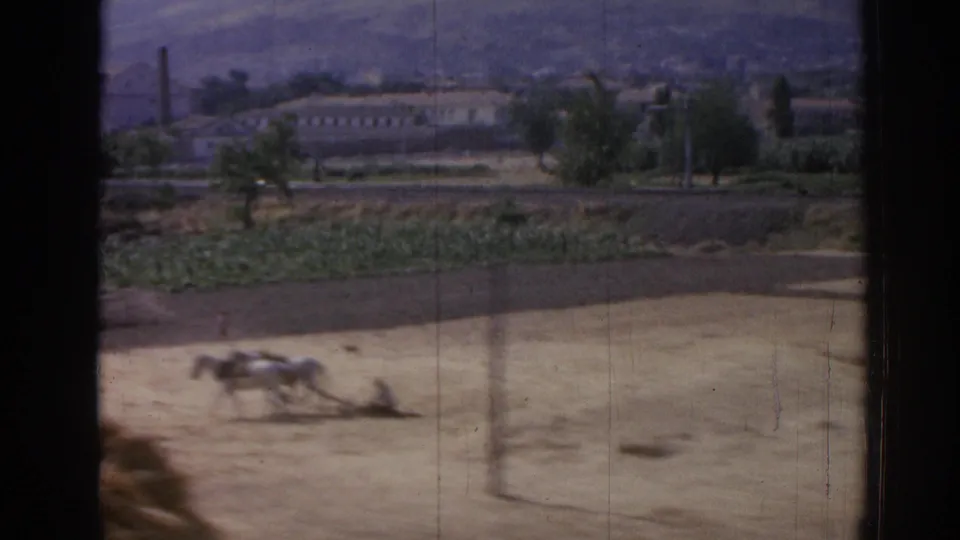 1962 Izmir-area home-movie frame showing a farmer with two horses plowing or crossing a dry field below distant buildings and hills.