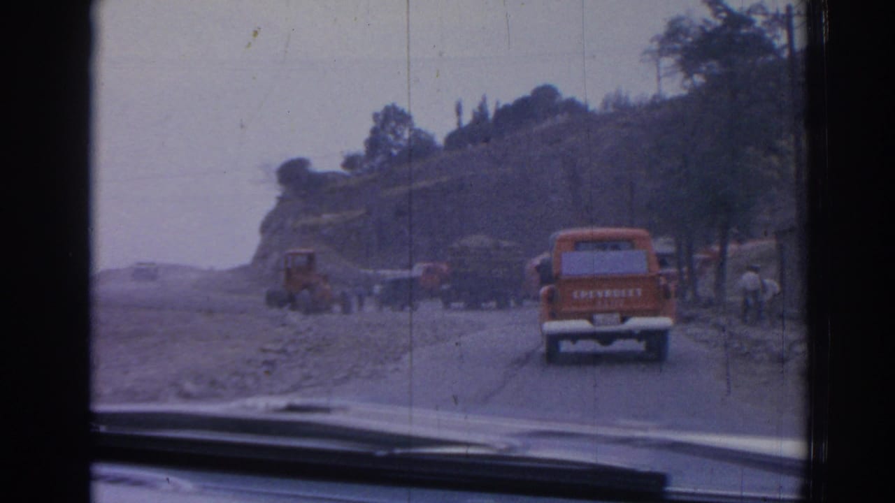 Orange Chevrolet truck drives along a dusty road past a tractor and other vehicles.