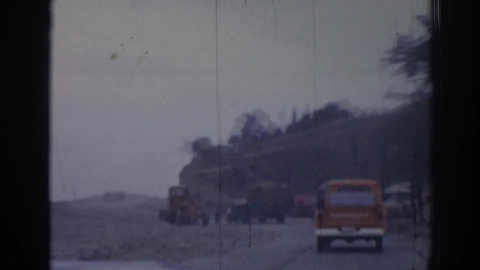 Soft 8mm frame from 1962 Izmir showing an orange truck, tractor, and roadside vehicles on a hazy rural road.