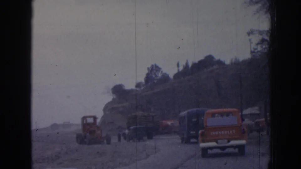 1962 Izmir home-movie frame with an orange Chevrolet truck, tractor, pedestrians, and parked vehicles along a rural hillside road.