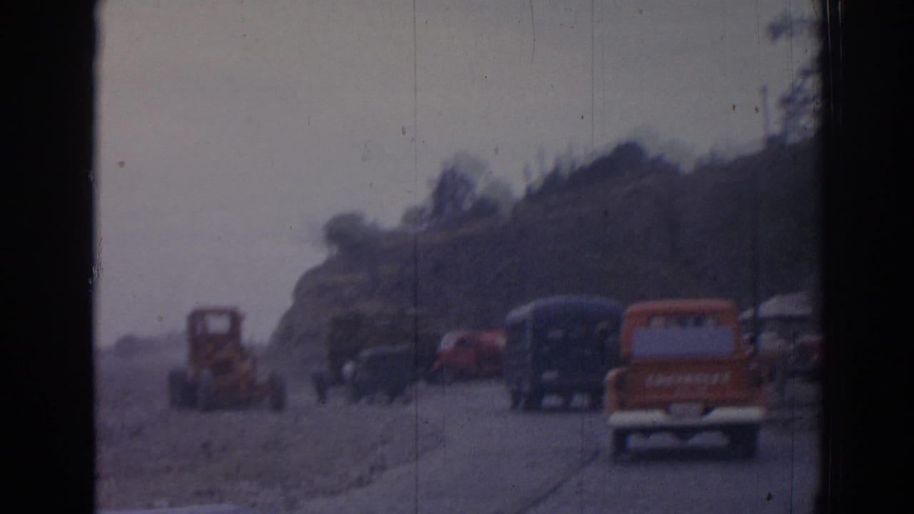 Vehicles travel on a dusty road with an orange tractor ahead and dark buses near a hillside.