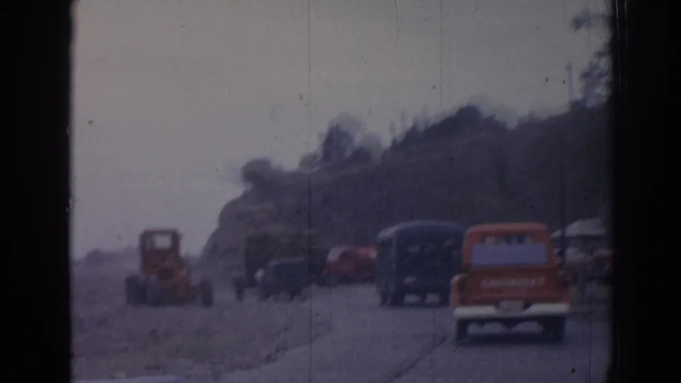 1962 Izmir home-movie frame showing an orange tractor, dark buses, and roadside traffic on a dusty rural road.