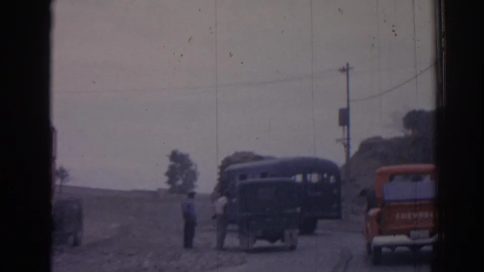 1962 Izmir-area roadside view with a dark military bus, red pickup, uniformed men, utility pole, and window-frame obstruction.