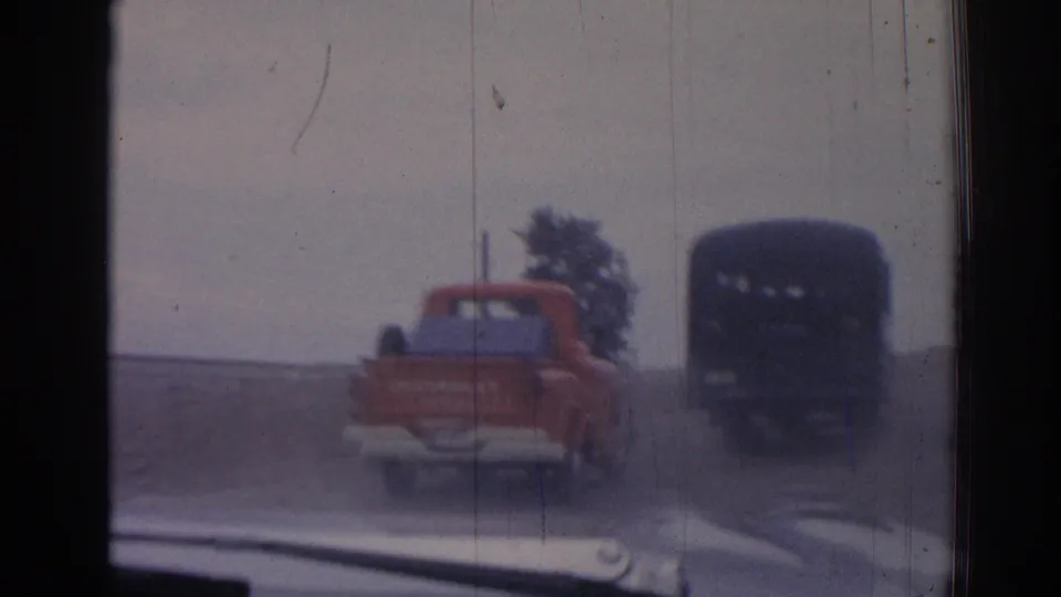In this 1962 Turkey home-movie frame, a red pickup follows a dark bus along a dusty rural road viewed through a window.