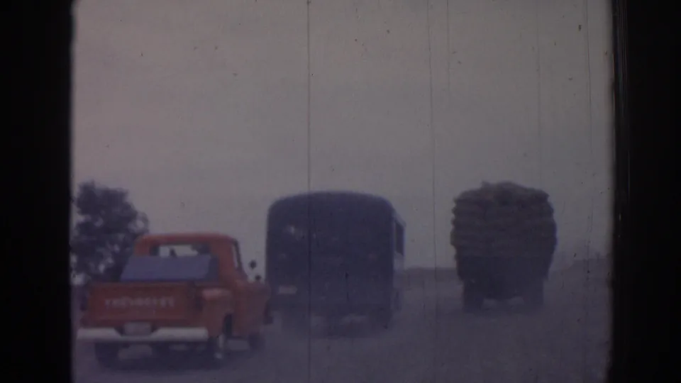Vehicles including a red pickup, dark bus, and loaded truck move away on a rural road outside Izmir in 1962 footage.