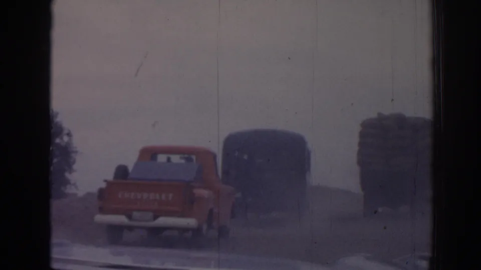 1962 Izmir frame with a red Chevrolet pickup leading dark trucks past a stacked cargo load in hazy light.
