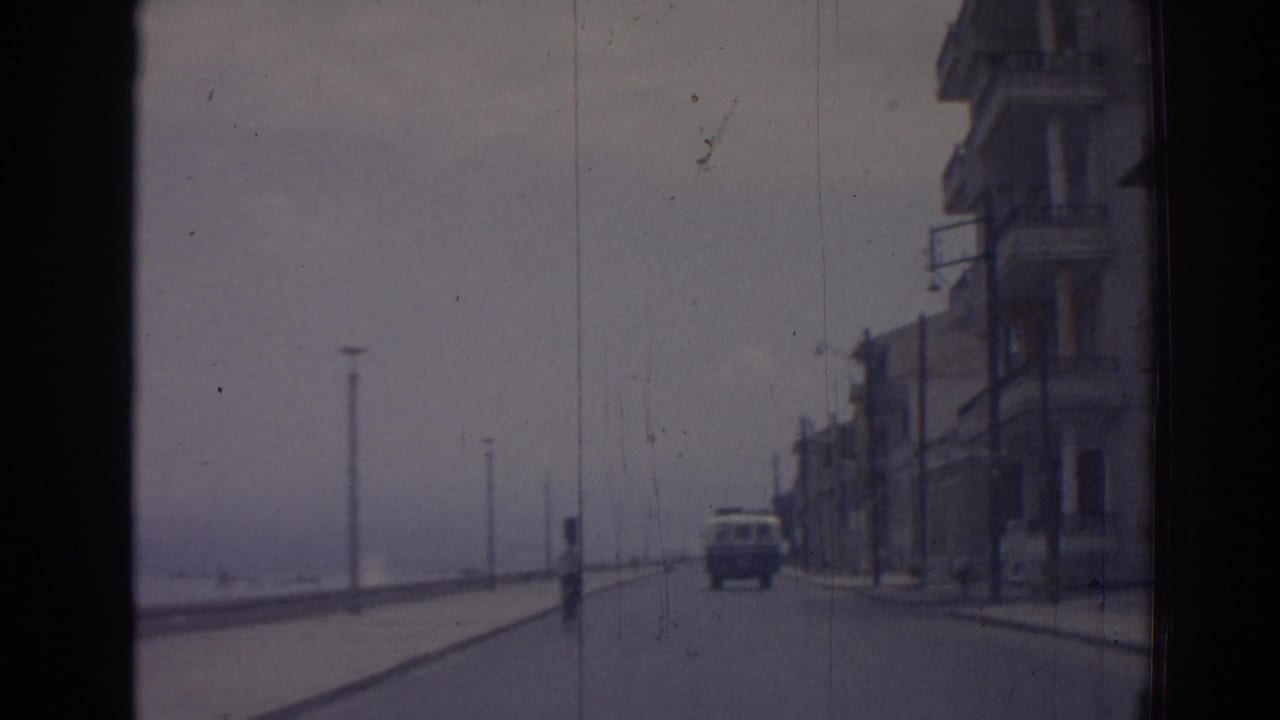 1962 Izmir home-movie frame from a moving car, showing a quiet waterfront road, sidewalk buildings, lampposts, and a vehicle ahead.