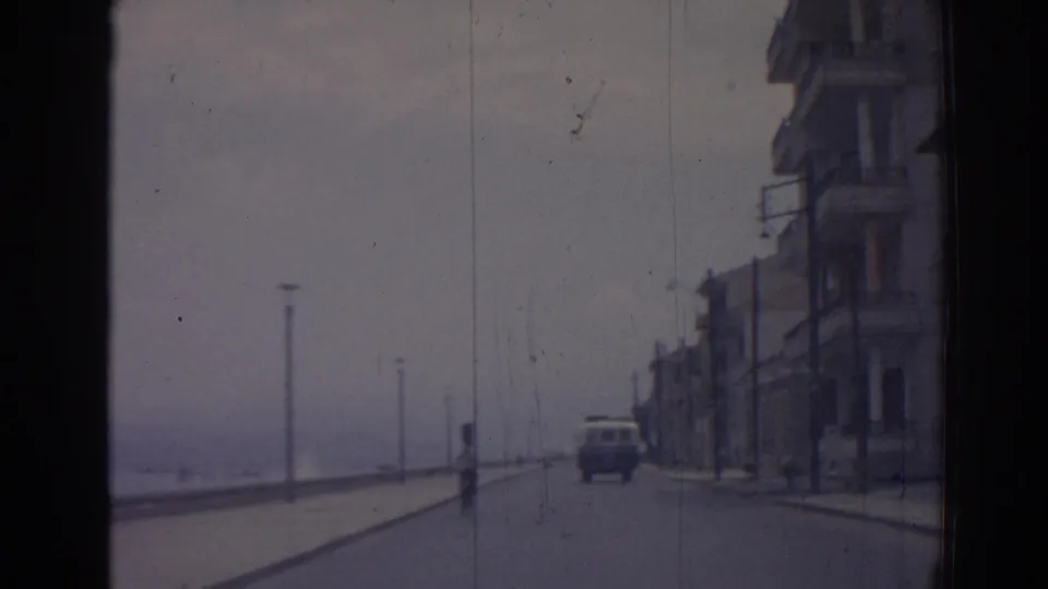1962 Izmir home-movie frame from a moving car, showing a quiet waterfront road, sidewalk buildings, lampposts, and a vehicle ahead.