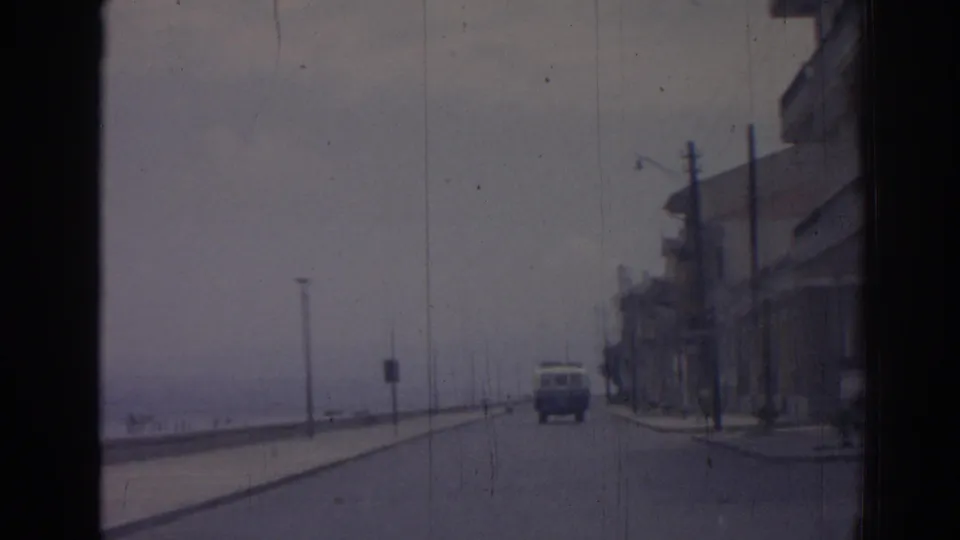 1962 Izmir street view from a moving car, with a white van ahead, sea to the left, lampposts, and multi-story buildings to the right.