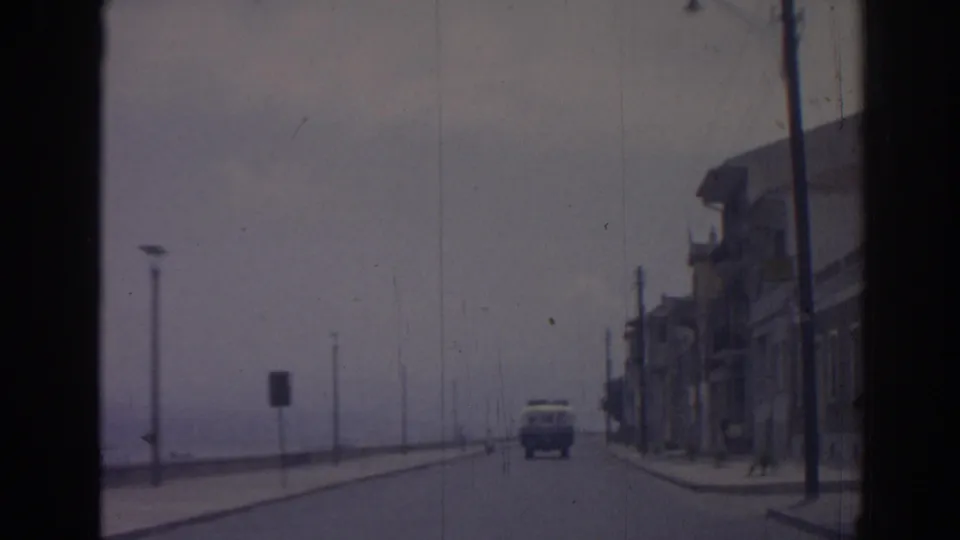 1962 Izmir coastal driving frame showing a van ahead, sparse street furniture, waterfront haze, buildings, and a distant person near the road.