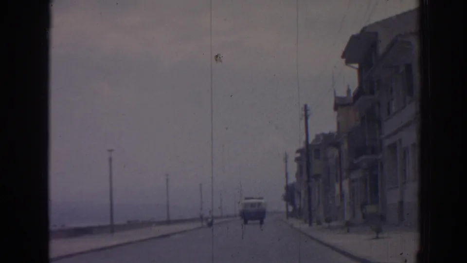 1962 Izmir home-movie frame from a car, showing a van on the coastal road, waterfront lamps, right-side buildings, and a distant person near the sidewalk.