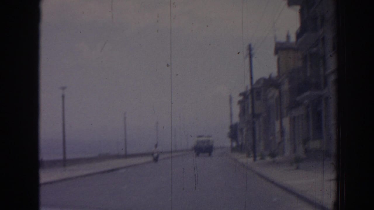 Coastal road with a scooter ahead, buildings on the right, and sea to the left.