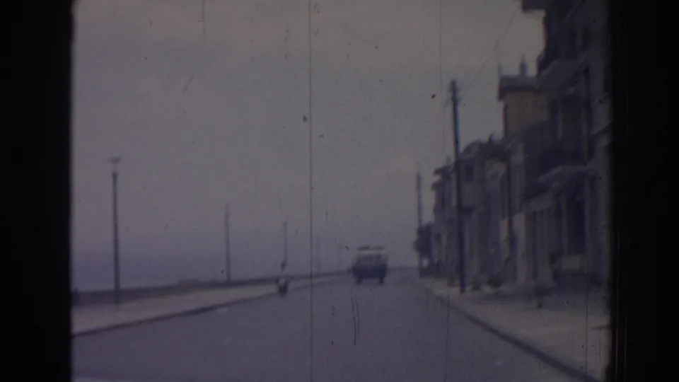 1962 Izmir coastal road seen from a moving vehicle, with a scooter ahead, roadside buildings, and the sea beyond the pavement.