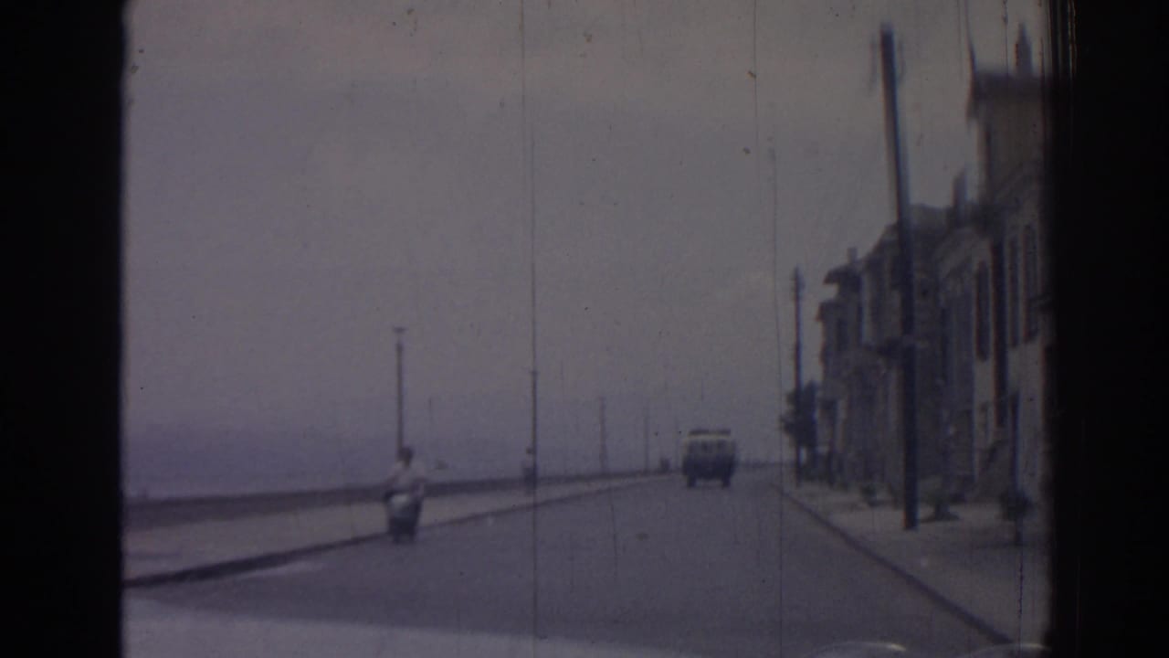 Vehicle approaches along a wide seafront road with buildings and a scooter nearby.