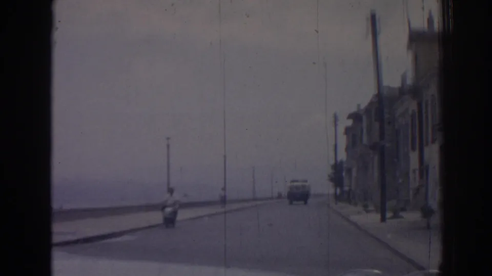 Moving 8mm view of an Izmir seafront road in 1962, with a scooter, a distant car, street lamps, and buildings lining the right side.