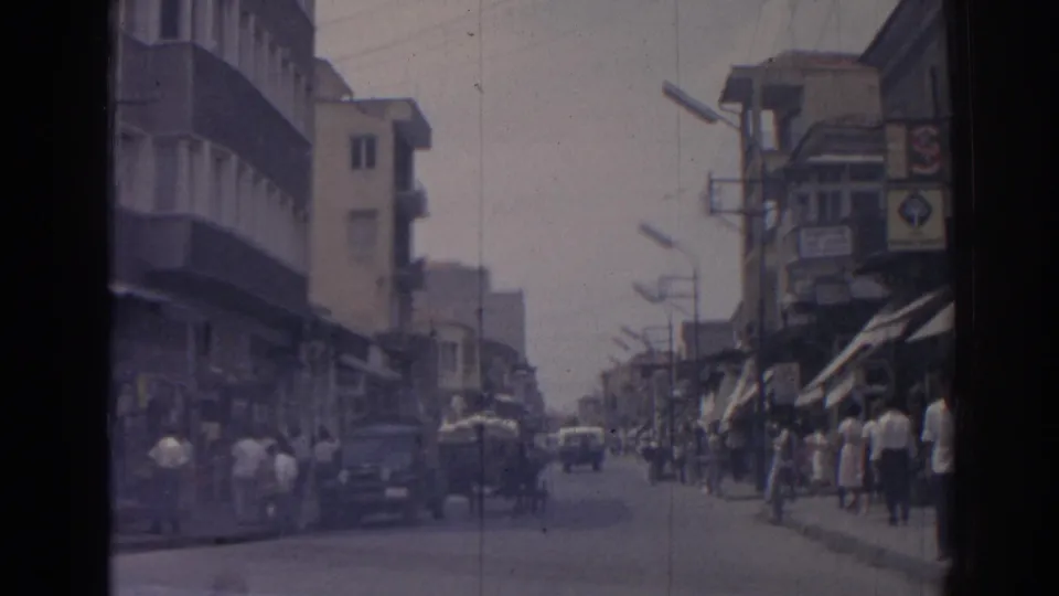 1962 Izmir street view with pedestrians near storefront awnings, classic cars, balconies, and a cart sharing the road.
