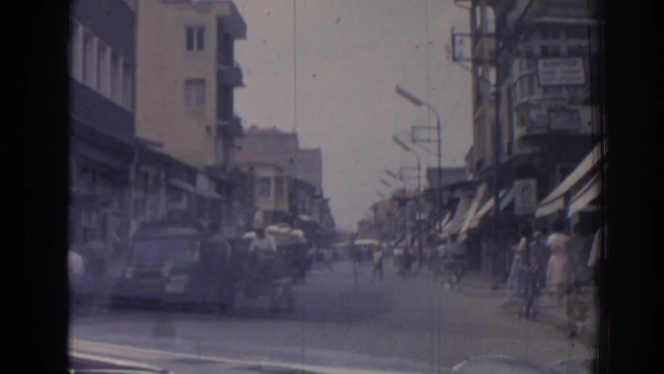 Soft 8mm frame of a 1962 Izmir commercial street with a passing car, pedestrians, bicycles, awnings, and balconies.