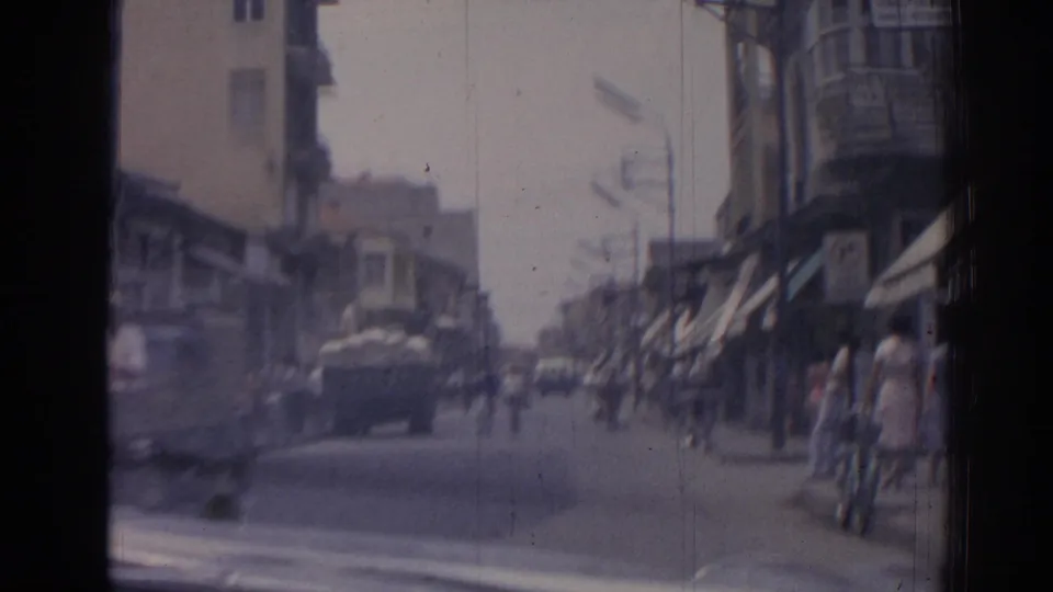 1962 Izmir street with pedestrians, cyclists, classic cars, awnings, balconies, and a loaded cart or truck visible ahead.