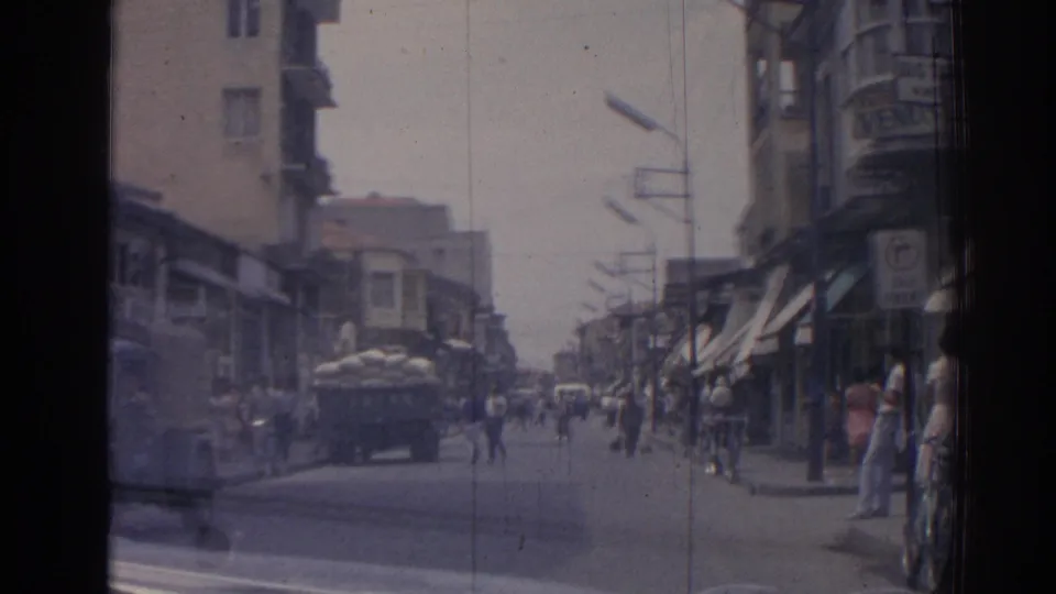 1962 Izmir street seen from a moving car, with pedestrians along storefronts and a sack-loaded truck in the road.