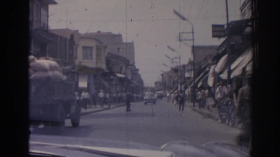 Moving 8mm view of a 1962 Izmir street, with pedestrians on sidewalks, a truck at left, and cars ahead.