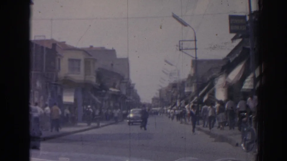 Late-frame view of a busy Izmir street in 1962, showing pedestrians, a car in the road, and storefront awnings.