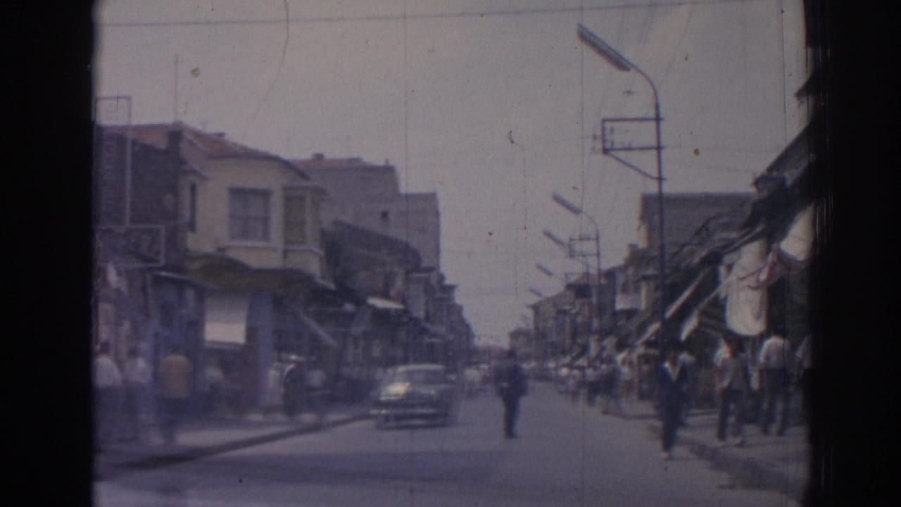 1962 Izmir street scene from a moving vehicle, with pedestrians on sidewalks and a classic car approaching near awning-covered shops.