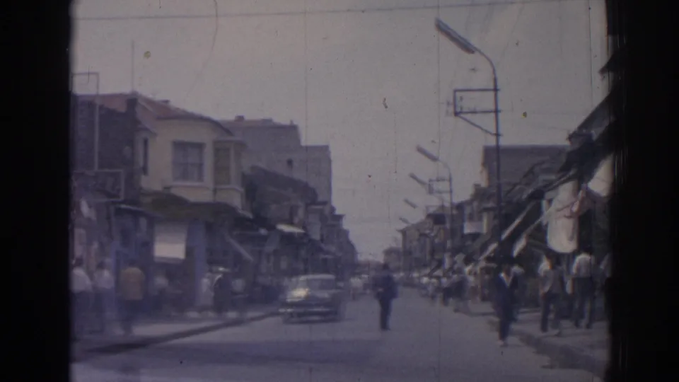 1962 Izmir street scene from a moving vehicle, with pedestrians on sidewalks and a classic car approaching near awning-covered shops.