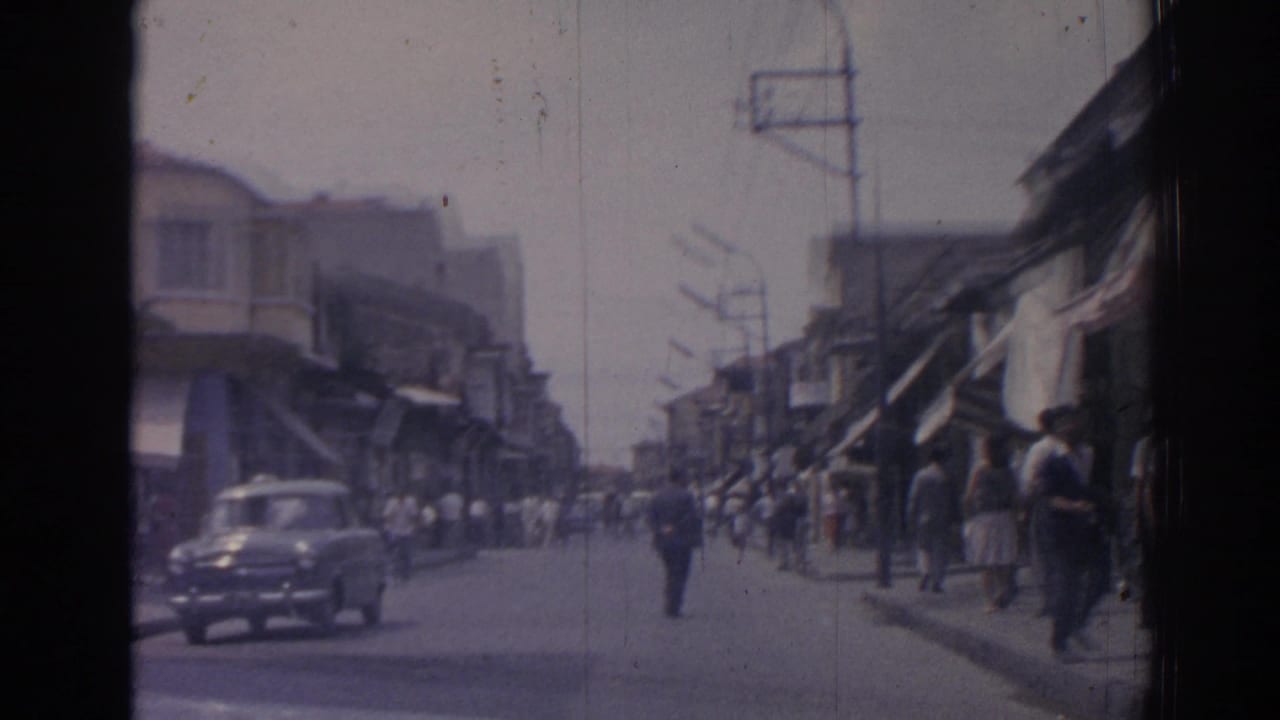 Classic car passes through a busy street lined with pedestrians and awnings.