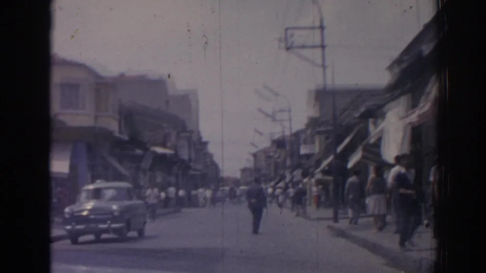 Moving view along a 1962 Izmir commercial street as a dark classic car passes and pedestrians stand along the sidewalks.
