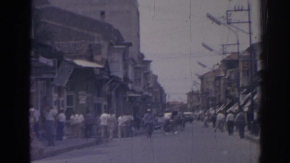 1962 Izmir street view from a moving vehicle, showing sidewalk crowds, awning-covered shops, overhead poles, and distant traffic.